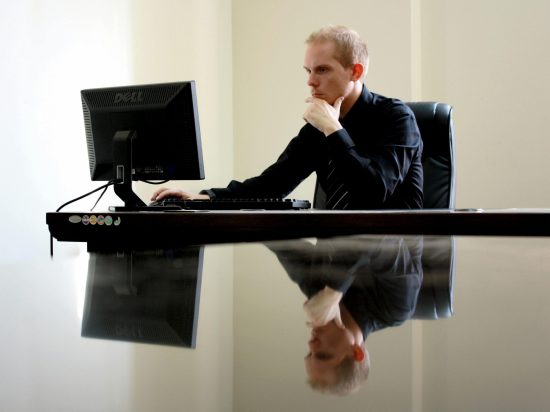 pexels-davidmceachan-90333 A man in a black dress shirt sits at a desk with his hand on his chin, looking intently at a computer monitor